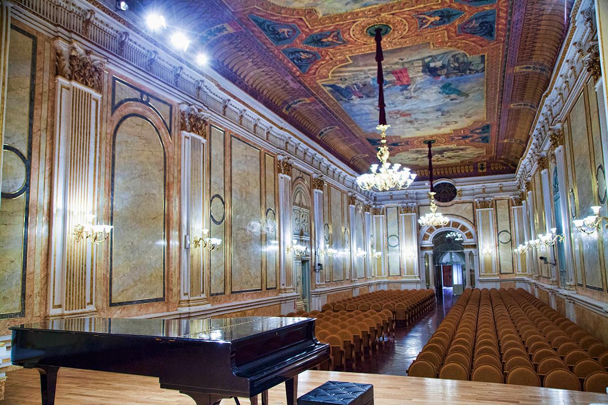 Interior de una sala de conciertos ornamentada con techo pintado, lámparas de araña, filas de butacas y un piano de cola en primer plano, en tonos cálidos dorados